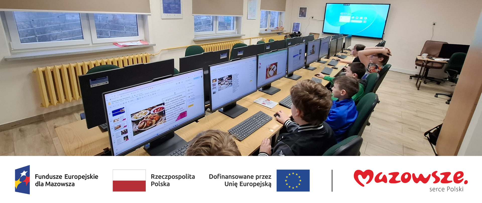 Wnętrze sali lekcyjnej, uczniowie siedzą przy komputerach, robią prezentację. Poniżej widoczne logo Funduszy Europejskich, Rzeczypospolitej Polskiej, Unii Europejskiej oraz województwa Mazowieckiego.