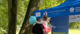 Mężczyzna trzymający na rękach dziecko odwiedza stoisko informacyjno-edukacyjne projektu LIFE Podkarpackie „Żyj i oddychaj” podczas wydarzenia plenerowego w parku. Stoisko z materiałami promocyjnymi i edukacyjnymi znajduje się pod niebieskim namiotem, w otoczeniu drzew i zieleni.