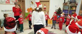 Dzieci ubrane na czerwono, w czapkach świętego Mikołaja tańczą w kółku, Panie nauczycielki bawią się razem z nimi