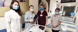Ostatni noworodek urodzony w 2020 roku w szpitalu powiatowym, leżący w łóżeczku na korytarzu oddziału ginekologiczno-położniczego. Obok szczęśliwa mama oraz personel medyczny. 