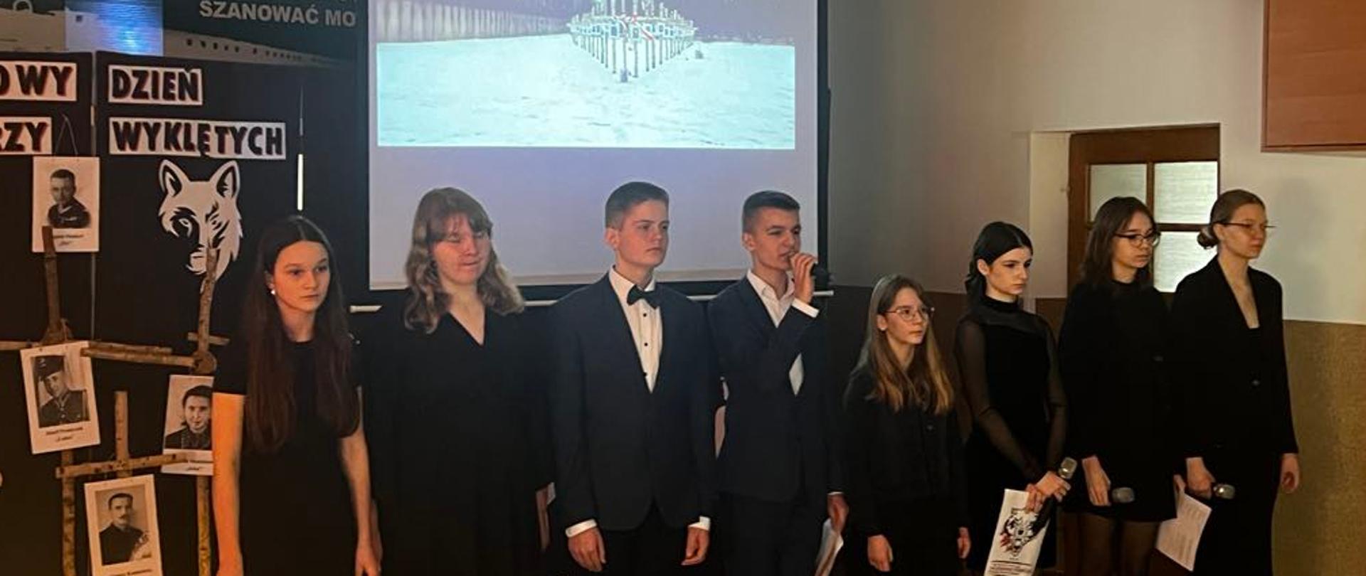 Na scenie stoi ośmioro uczniów ubranych na czarno. Chłopiec stojący w środku przemawia przez mikrofon. Za nimi znajduje się scenografia - brzozowe krzyże, zdjęcia żołnierzy i napis: 1 MARCA - NARODOWY DZIEŃ PAMIĘCI ŻOŁNIERZY WYKLĘTYCH. Na podłodze stoją zapalone znicze.