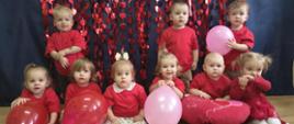 Na zdjęciu są dzieci z grupy Pszczółki. Dzieci ubrane są w czerwone koszulki. W tle jest "serduszkowa" dekoracja. Część dzieci trzyma w rączkach czerwone lub różowe balony.
