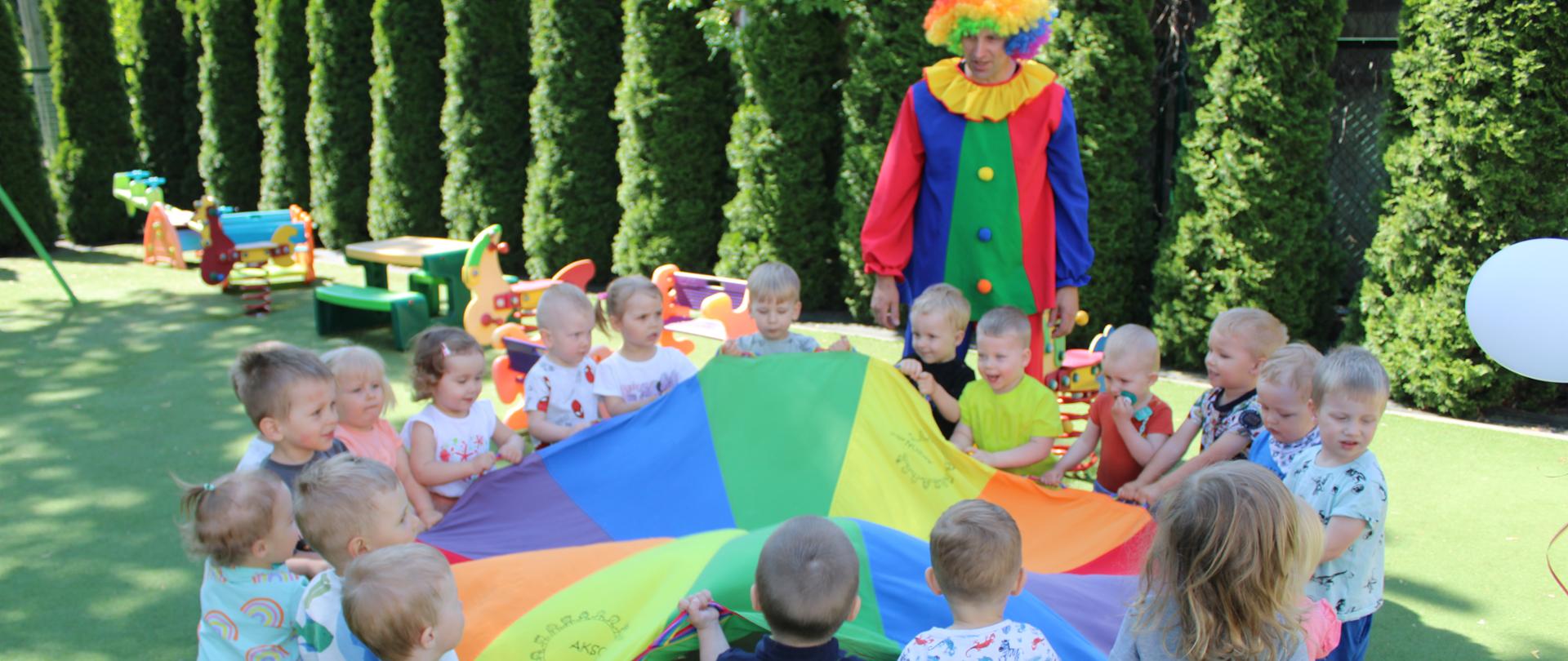Dzieci z grupy Motylki stoją w kole, trzymająć kolorową chustę animacyną. W tle widać Klauna.
