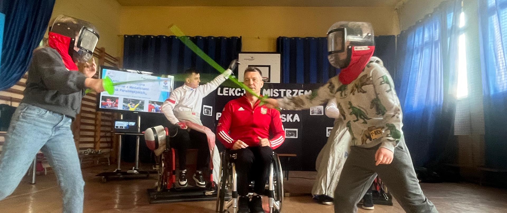 Dwoje dzieci w maskach sportowych trzyma prawurącz zielone szpady ku sobie. W tle siedzi mężczyzna w czerwonej koszulce na wózku inwalidzkim