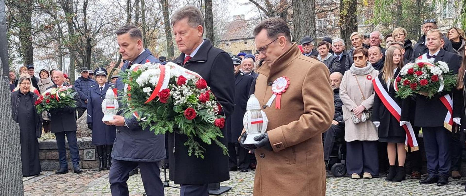 Na fotografii widzimy od lewej wicestarostę Zbigniewa Czerkasa, przewodniczącego Rady Powiatu Sokołowskiego Jarosława Puściona oraz Starostę Sokołowskiego Adama Górala. Przewodniczący niesie w dłoniach wiązankę patriotyczną z biało - czerwonymi szarfami, natomiast Wicestarosta i Starosta znicze z biało - czerwoną flagą. Delegacja zbliża się do czoła pomnika "Czynu Niepodległościowego' w Sokołowie Podlaskim, pod którym za chwilę złoży kwiaty i znicze. W tle pozostałe delegacje stojące w kręgu oraz uczestnicy uroczystości.