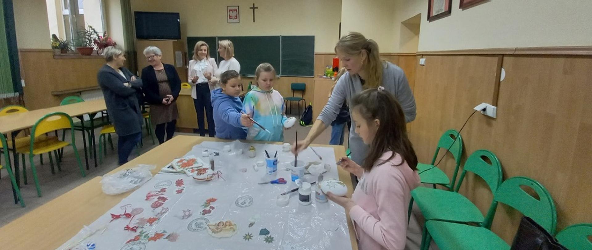 troje dzieci i dorosła kobieta stoją wokół dużego stołu i malują farbami. z tyły cztery kobiety rozmawiają ze sobą obserwując dzieci 
