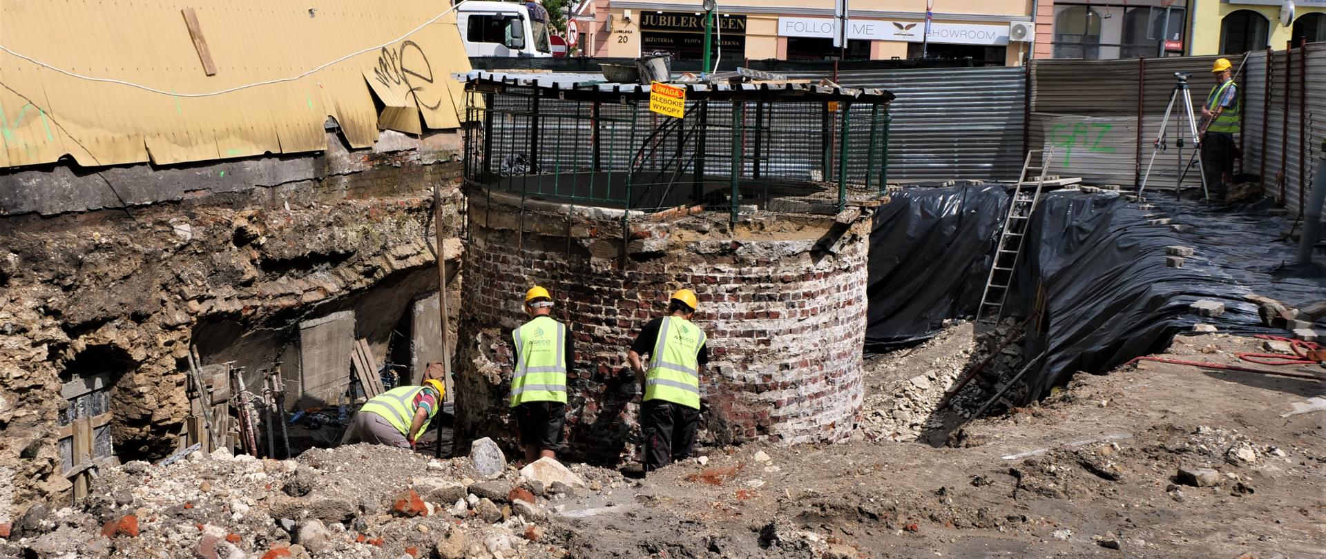 Zdjęcie przedstawia trzy osoby pracujące w wykopanym dole na placu budowy na zewnątrz