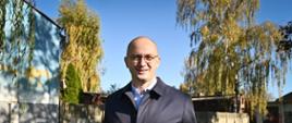 Mężczyzna w okularach stoi na trawie i trzyma tablicę z napisem: ‘Zazielenimy Konstantynów drzewami, kwiatami, bylinami’. W tle zieleń i budynki.