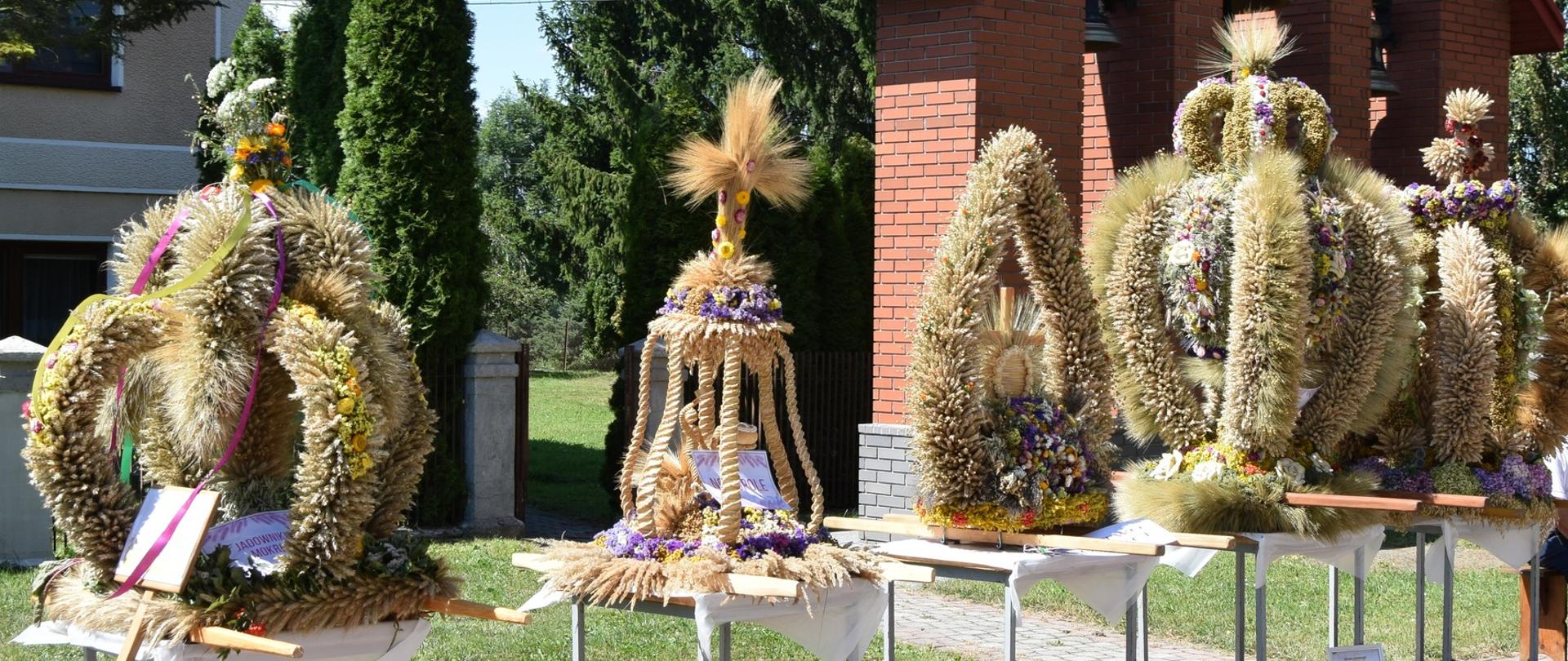 Tradycyjne wieńce dożynkowe wykonane z kłosów zbóż, kwiatów i wstążek, ustawione na stołach przed budynkiem podczas święta plonów. W tle dwóch mężczyzn siedzi na ławce przy czerwonej kapliczce.