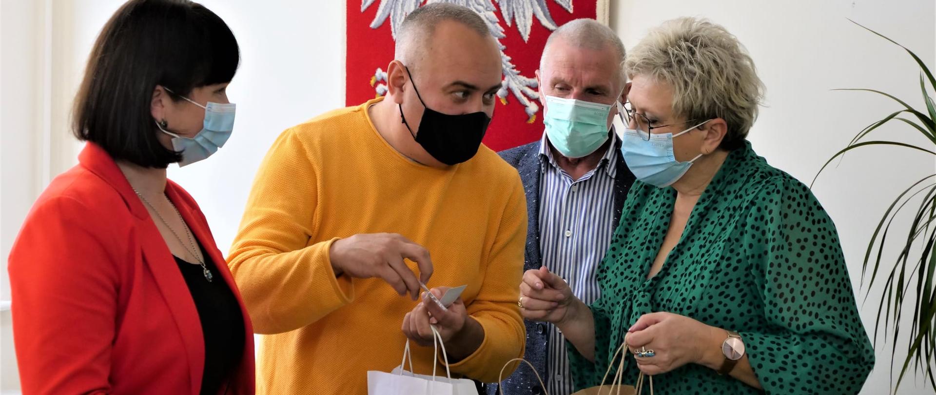 Cztery osoby stoją wewnątrz pokoju na tle ściany. Trzymają w rękach reklamówki z podarunkami. Rozmawiają. Mają na twarzach maseczki.
