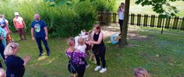 Festyn Profilaktyczno Zdrowotny w ogrodach przasnyskiego szpitala. Zdjęcie dziewczynkę w rękach matki, karmiącą alpakę pod czujnym okiem trenerki zwierząt. Po lewej stronie pozostali uczestnicy Festynu oraz Starosta Przasnyski Krzysztof Bieńkowski.