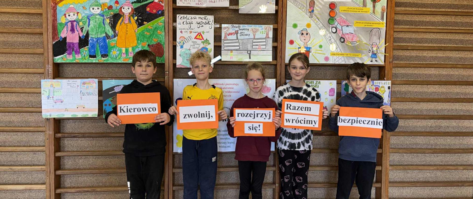 Pięcioro uczniów - dwie dziewczynki i trzech chłopców trzymają pomarańczowe kartki z przyklejonymi do nich słowami. Słowa na kartkach tworzą napis: ,,Kierowco zwolnij, rozejrzyj się! Razem wrócimy bezpiecznie". Z tyłu za dziećmi wiszą plakaty promujące akcję ,,Odblaskowa Szkoła".