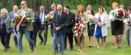 Uczestnicy wydarzenia idą po zielonej łące na tle lasu niosąc w dłoniach wiązanki.