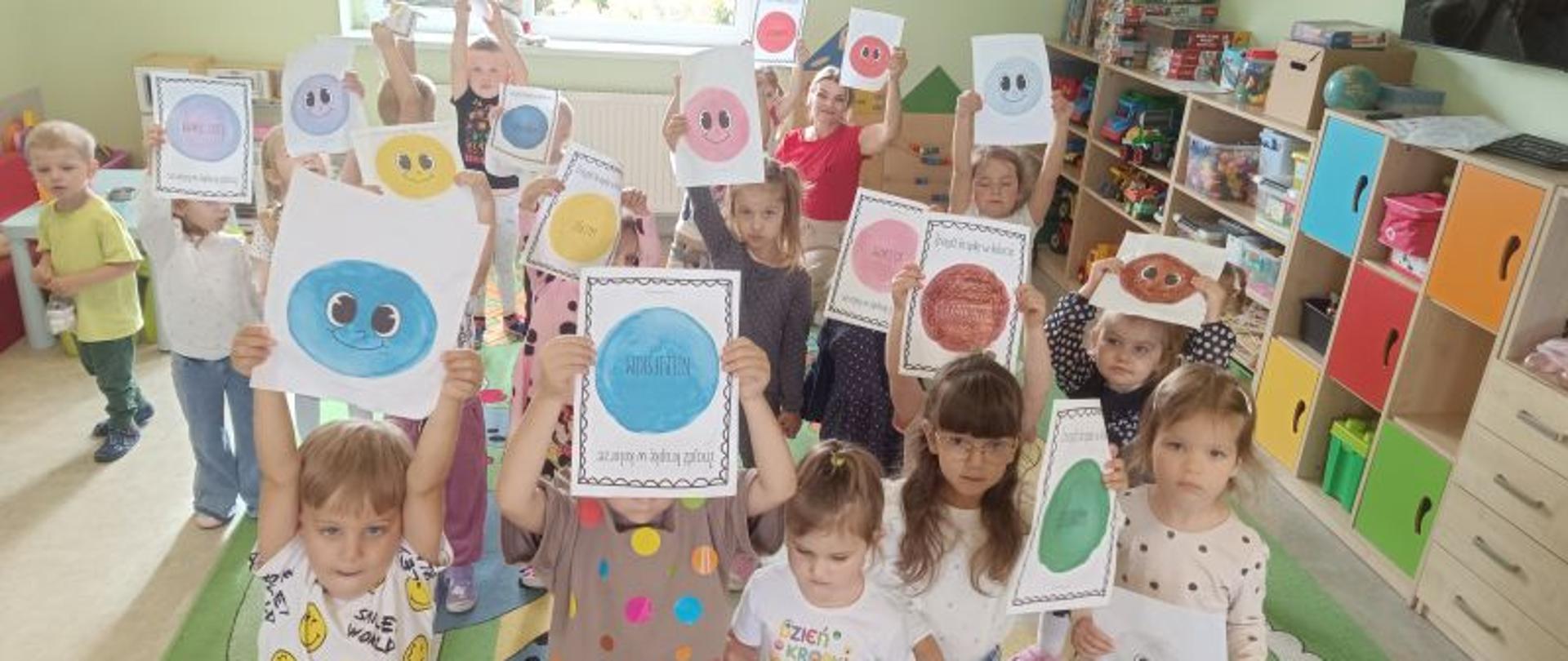 Dzieci stoją na dywanie i pokazują swoje prace plastyczne wykonane z okazji Dnia Kropki.