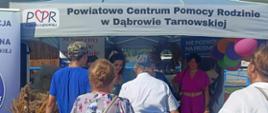 Zdjęcie przestawia stoisko Powiatowego Centrum Pomocy Rodzinie w Dąbrowie Tarnowskiej w trakcie trwania Dożynek Powiatowych w 2024 roku. Przed stoiskiem widać osoby zainteresowane materiałami promocyjnymi.