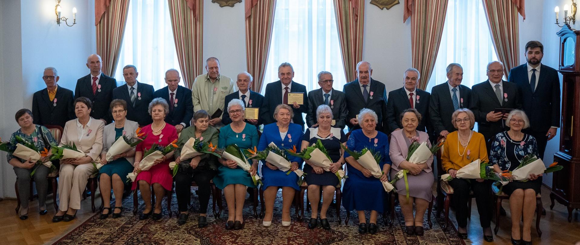 Zdjęcie przedstawia 25 osób wewnątrz budynku. W pierwszym rzędzie kobiety siedzą na krzesłach, a za nimi stoją mężczyźni. Kobiety trzymają w rękach bukiety kwiatów.