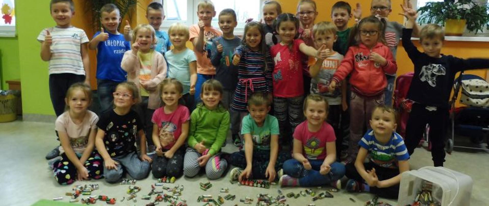 Grupa dzieci siedzi lub stoi, przed nimi leżą baterie. W tle dwa okna, na szybach widać kolorowe liście. 