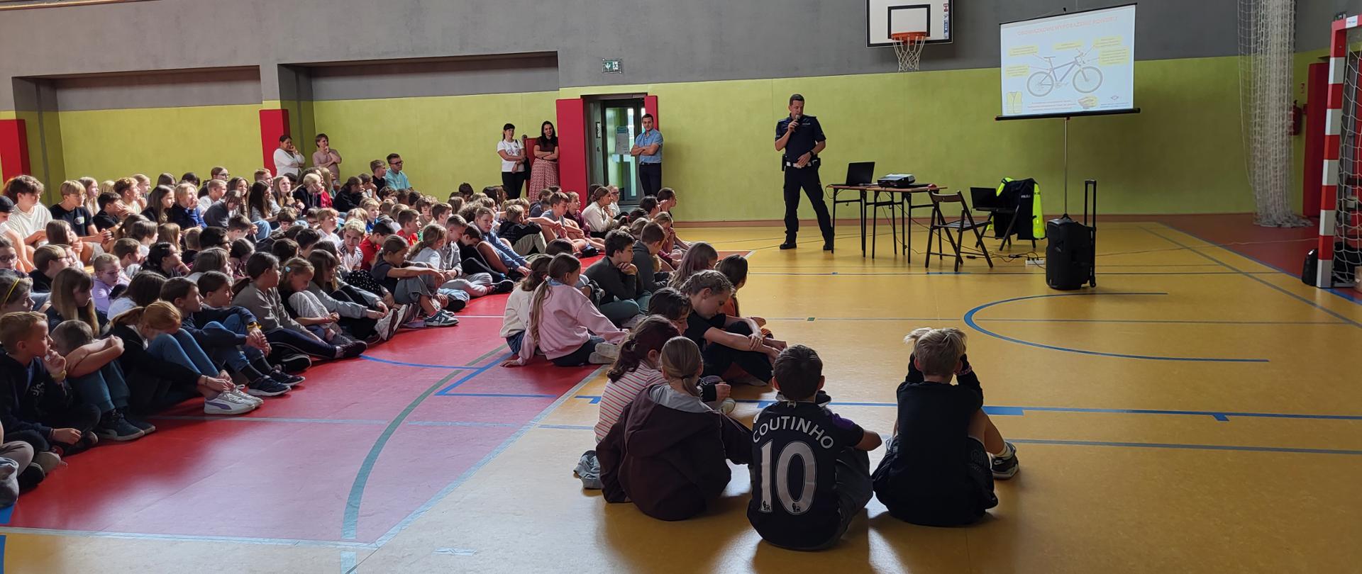 Grupa uczniów siedzi na sali gimnastycznej i słucha przemawiającego policjanta