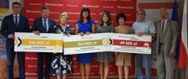 Osiem osób stoi na tle czerwonej ścianki z napisami Mazowsze. Trzymają trzy duże kartony z kwotami 300000 zł, 164000 zł oraz 49500 zł