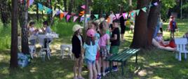 Dzieci stojące w grupie, siedzące przy białym stoliku i leżące na kocu wśród drzew, nad mini rozciągają się kolorowe chorągiewki