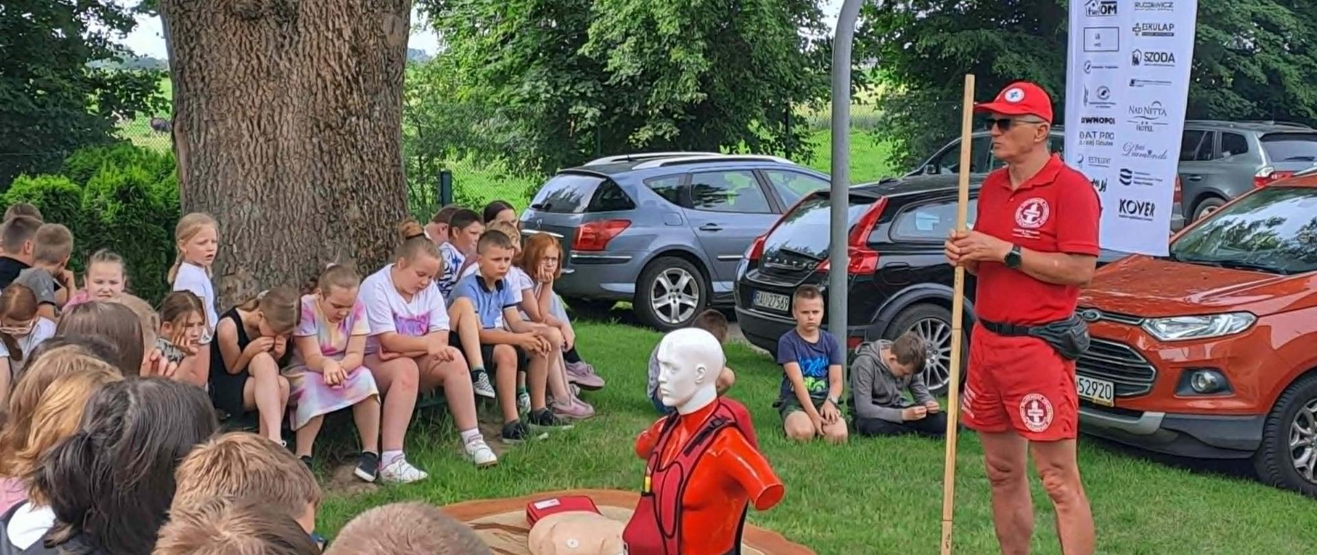 Zdjęcie przedstawia pokaz pierwszej pomocy prowadzony przez ratownika dla grupy dzieci, zorganizowany na świeżym powietrzu pod dużym drzewem.