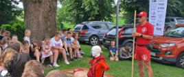 Zdjęcie przedstawia pokaz pierwszej pomocy prowadzony przez ratownika dla grupy dzieci, zorganizowany na świeżym powietrzu pod dużym drzewem.