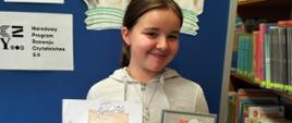 Dziewczynka z klasy 5 trzyma w jednej ręce wypożyczoną z biblioteki książkę przedstawiającą domek jednorodzinny i chłopca obok, w drugiej ręce wykonany przez siebie obrazek z takim samym motywem