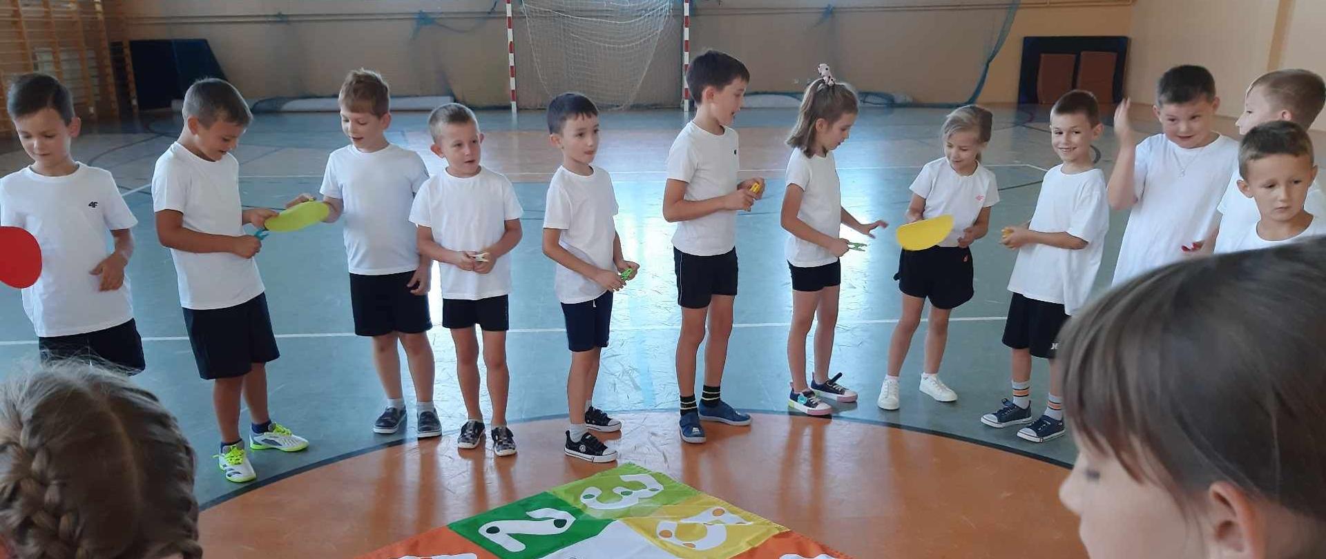 Uczniowie w stroju sportowym na hali gimnastycznej, trzymaj w rękach spinacze i przekładaj koło bo obwodzie koło.