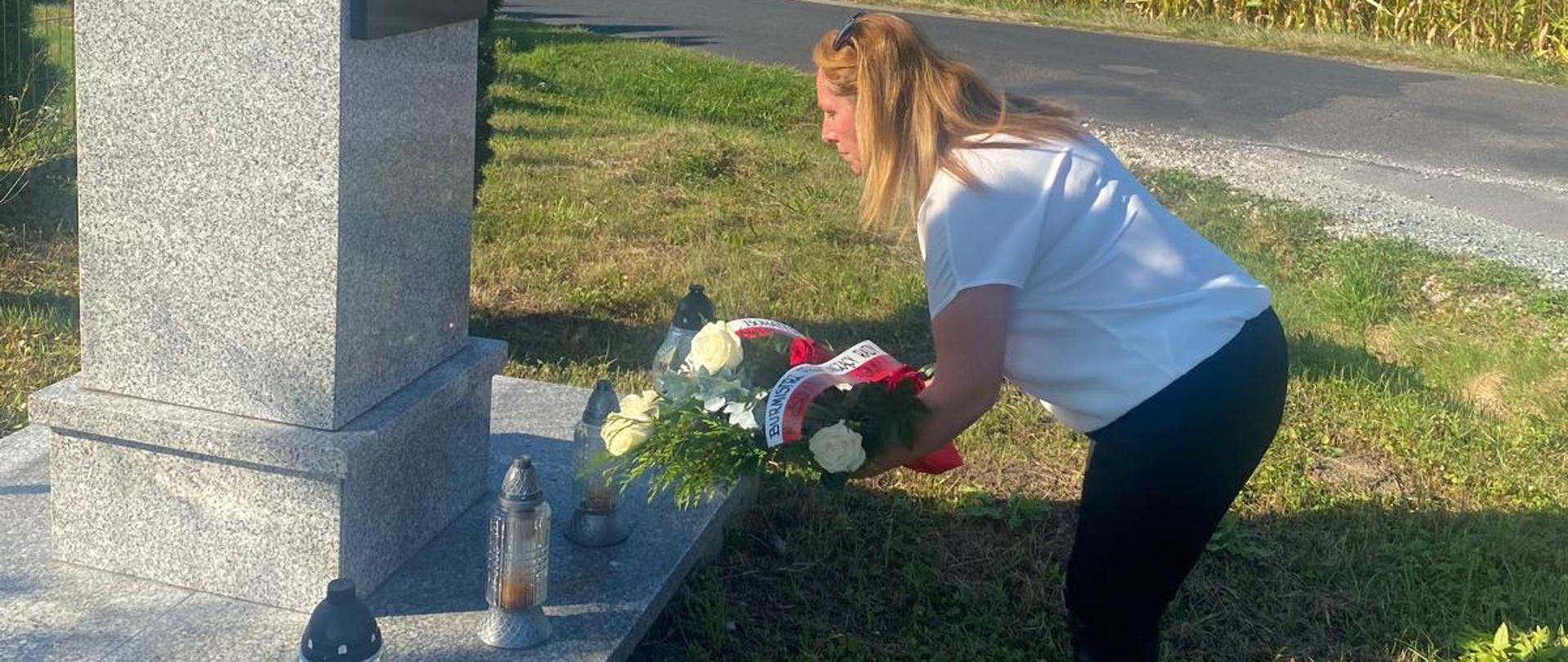 Kobieta w białej koszuli i czarnych spodniach składa wieniec pod pomnikiem. W tle wiejski krajobraz: zielona trawa, asfaltowa droga, niebieskie niebo.