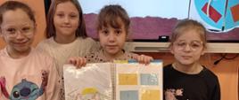 Cztery dziewczynki trzymają w ręku otwarty lapbook, za nimi na monitorze wyświetlona prezentacja o lapbooku