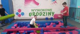 Jeden uczeń stoi w basenie z miękkimi gąbkowymi klockami, drugi uczeń stoi na mostku ponad nim i trzyma w ręku gąbkową belkę