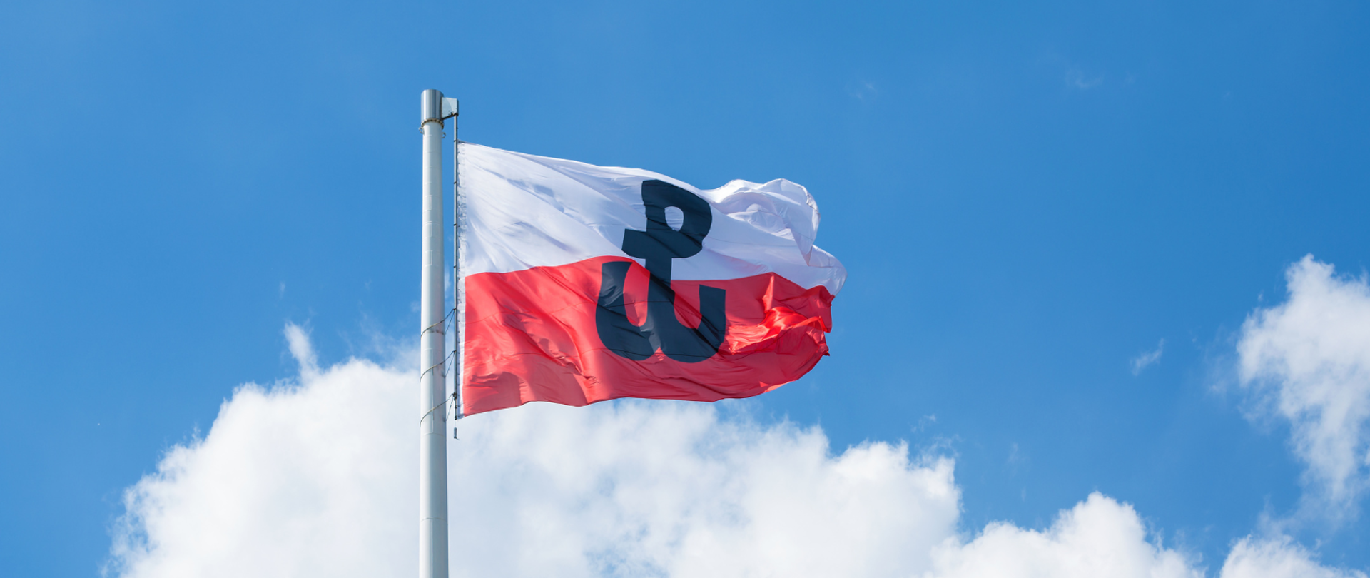 Flaga Polski z symbolem Polski Walczącej. Ta flaga składa się z dwóch poziomych pasów: białego na górze i czerwonego na dole, z czarnym symbolem kotwicy na środku. Powiewa ona na tle nieba o jasnym błękicie z kilkoma rozproszonymi chmurami.