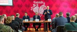 Zdjęcie przedstawia grupę ludzi podczas spotkania. Na pierwszym planie siedzący na krzesłach przedstawiciele Ochotniczych Stażu Pożarnych zwróceni w stronę stołu prezydialnego. Drugi plan stanowi stół prezydialny, za którym zwróceni w stronę słuchaczy znajdują się przedstawiciele władz powiatowych i wojewódzkich. W tle za nimi znajduje się czerwona ścianka promocyjna powiatu kraśnickiego z białym logo.