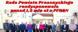 Grafika promująca informację o rozdysponowaniu przez Radnych Radny Powiatu Przasnyskiego ponad miliona trzystu tysięcy złotych z Państwowego Funduszu Rehabilitacji Osób Niepełnosprawnych. Na zdjęciu Radni Rady Powiatu Przasnyskiego stoją na dziedzińcu Starostwa Powiatowego w Przasnyszu przed wejściem do siedziby. 