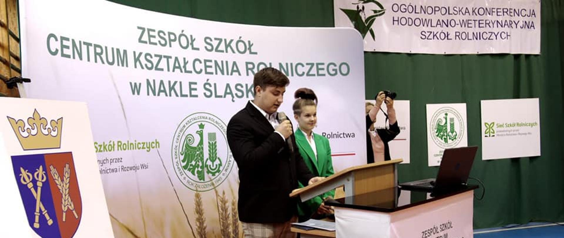 Konferencja w ZS CKR w Nakle Śląskim - przywitanie gości przez konferansjerów. W tle banner szkoły oraz Uniwersytetu Rolniczego w Krakowie