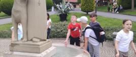 Dzieci wrzucają pieniążki do fontanny ozdobionej kamienną figurą zakonnika z psem