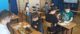 Dzieci siedzące przy stolikach. Pomiędzy nimi leżą szachownice
