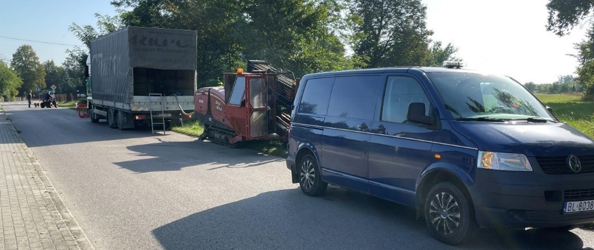 Droga z chodnikiem, na poboczu stoi granatowy bus. Za nim maszyna do robienia przewiertów oraz ciężarówka.
