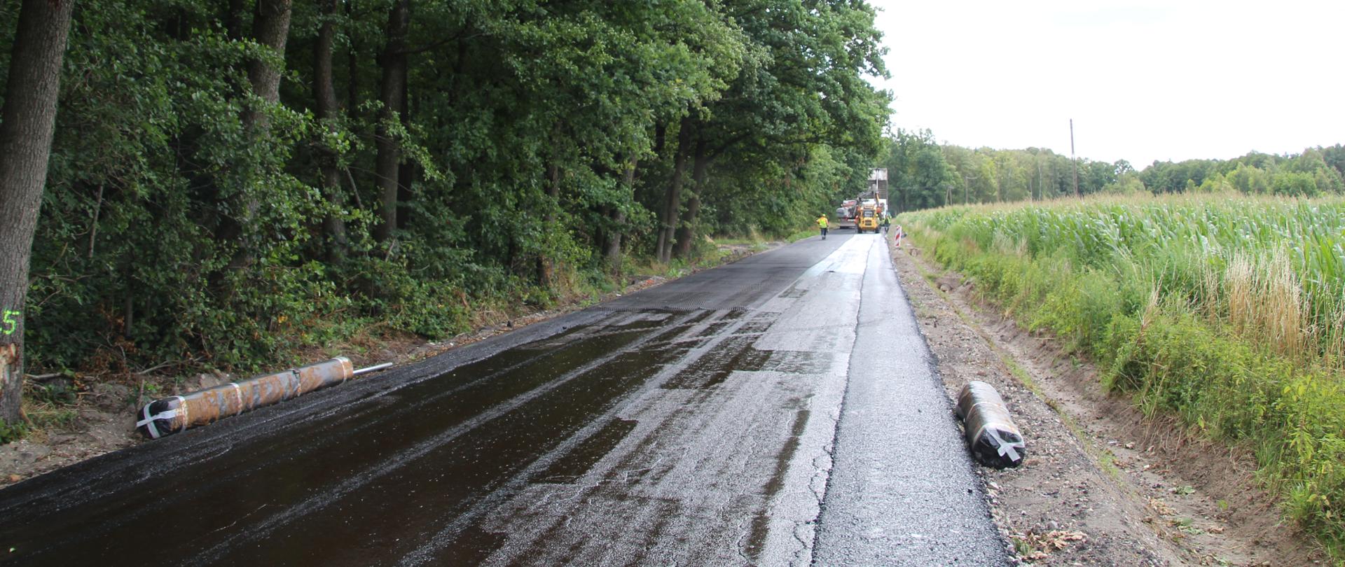 Trwa układanie nowej nawierzchni asfaltowej na drodze biegnącej wzdłuż lasu. Po lewej stronie widać gęste drzewa, po prawej pole kukurydzy. W głębi kadru pracują maszyny budowlane oraz robotnicy drogowi.