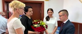 Kobieta w czarnej bluzce wręcza duży bukiet czerwonych róż i zielonych kwiatów mężczyźnie w ciemnym garniturze i okularach. Obok stoją dwie inne kobiety – jedna w jasnej bluzce z bliska, druga w białej sukience w tle. Spotkanie odbywa się w eleganckim pomieszczeniu.