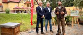 Osoby stoją na tle budynku i przemawiają do mikrofonu