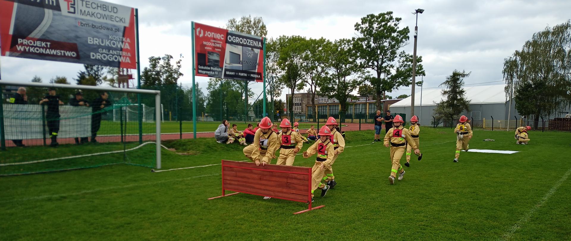 Na zdjęciu widzimy biegnących po murawie stadionu młodych strażaków, którzy przeskakują przez płotek stanowiący przeszkodę terenową. Strażacy ubrani w mundury bojowe oraz czerwone hełmy.