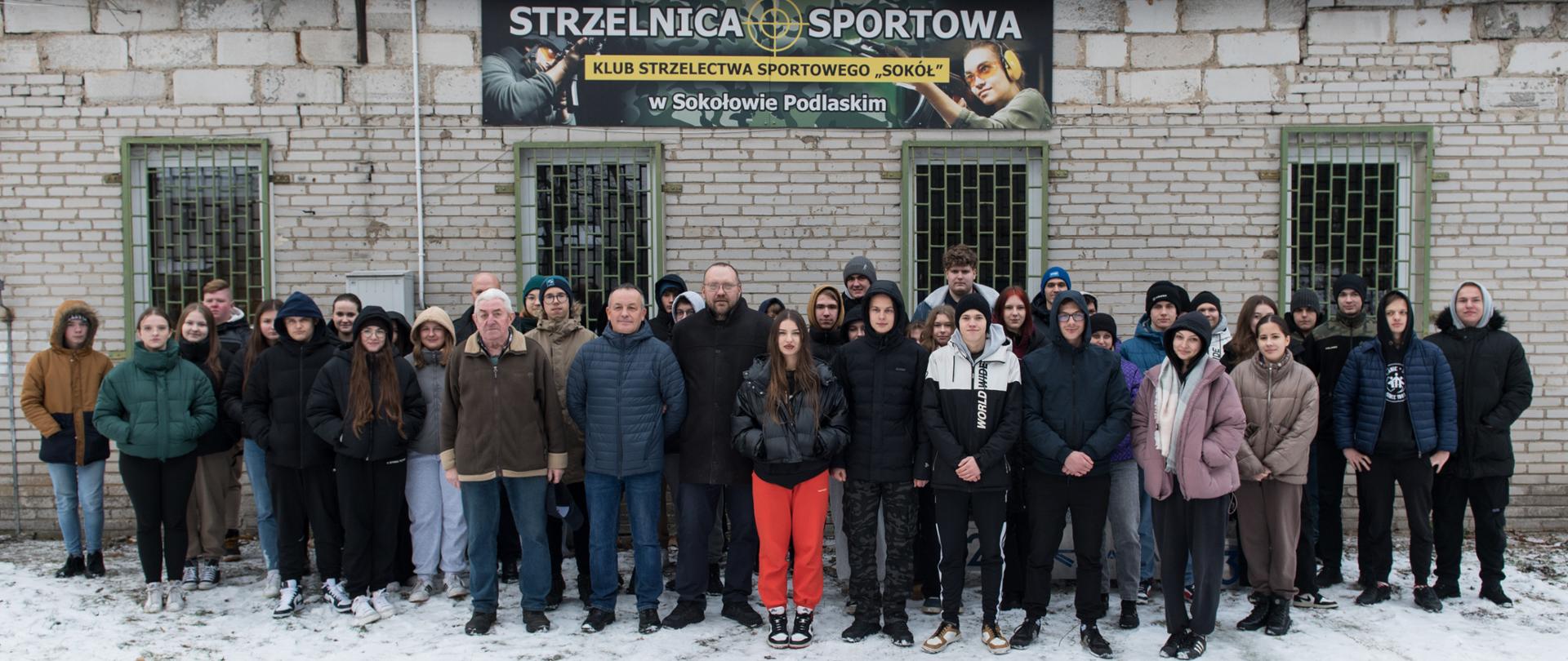 Fotografia grupowa uczestników turnieju strzeleckiego wraz z organizatorami na tle baneru Strzelnica Sportowa.