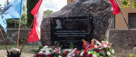 Na fotografii widzimy głaz do którego przymocowana została czarna prostokątna kamienna tablica. Na tablicy widnieje napis: "Romanowi Dmowskiemu, jednemu z Ojców Polskiej Niepodległości, którego korzenie rodzinne sięgają Dmoch w obecnej Gminie Bielany. W hołdzie społeczeństwo Gminy Bielany, Parafii Rozbity Kamień i Powiatu Sokołowskiego. Rozbity Kamień, Październik 2023 r. Po lewej i prawej strony tablicy widoczne: herb gminy Bielany oraz logo Powiatu Sokołowskiego. Na górze tablicy po lewej stronie wygrawerowano cytat z Romana Dmowskiego "Jestem Polakiem - więc mam obowiązki polskie". W lewym górnym rogu wygrawerowano wizerunek Romana Dmowskiego, a pod cytatem widoczny jest kontur granic gminy Bielany wraz z zaznaczonymi miejscowościami: Bielany, Rozbity Kamień oraz dawne Dmochy Mingosy. Pod pomnikiem złożone wiązanki z biało - czerwonych kwiatów, a po bokach flagi biało - czerwone oraz flaga z herbem gminy Bielany.
