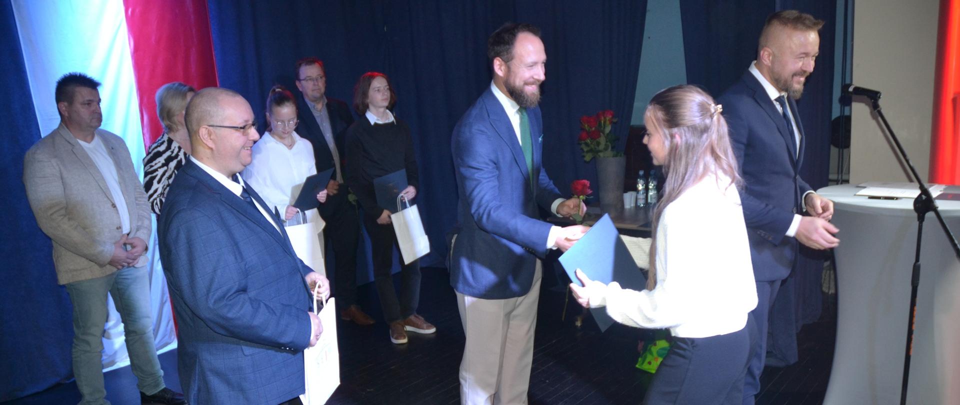Mężczyzna podaje rękę uczennicy oraz wręcza jej czerwoną różę. Obok stoją elegancko ubrani inni ludzie.