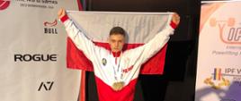 Odznaczony medalami Jakub Bońkowski pozuje z biało czerwoną flagą Polski. Flagę trzyma rozpostartą dwoma rękami za głową.