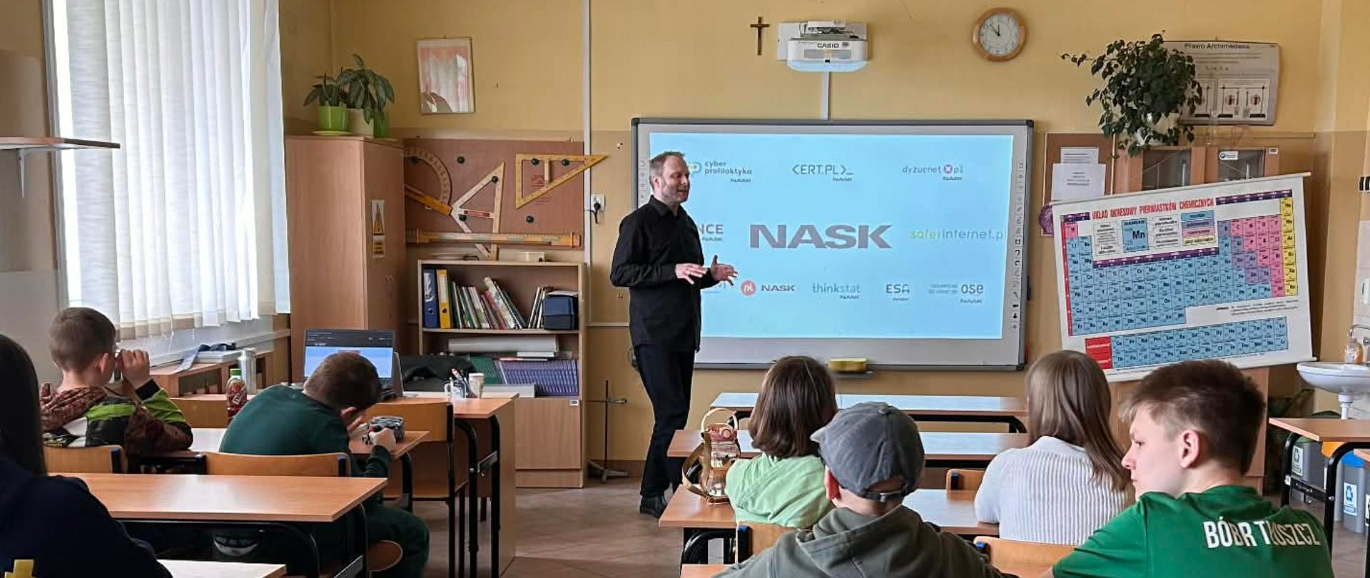 Mężczyzna w czarnej koszuli stoi przed uczniami siedzącymi w ławkach szkolnych. Na dużym ekranie na ścianie wyświetla się prezentacja z tytułem NASK