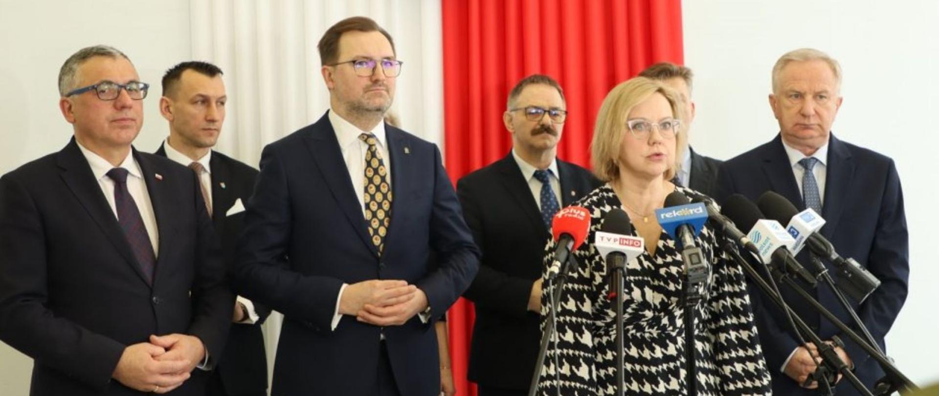 Minister Klimatu i Środowiska Anna Moskwa przemawia do zebranych, za nią stoją samorządowcy z województwa mazowieckiego. W tle ściana w barwach narodowych.
