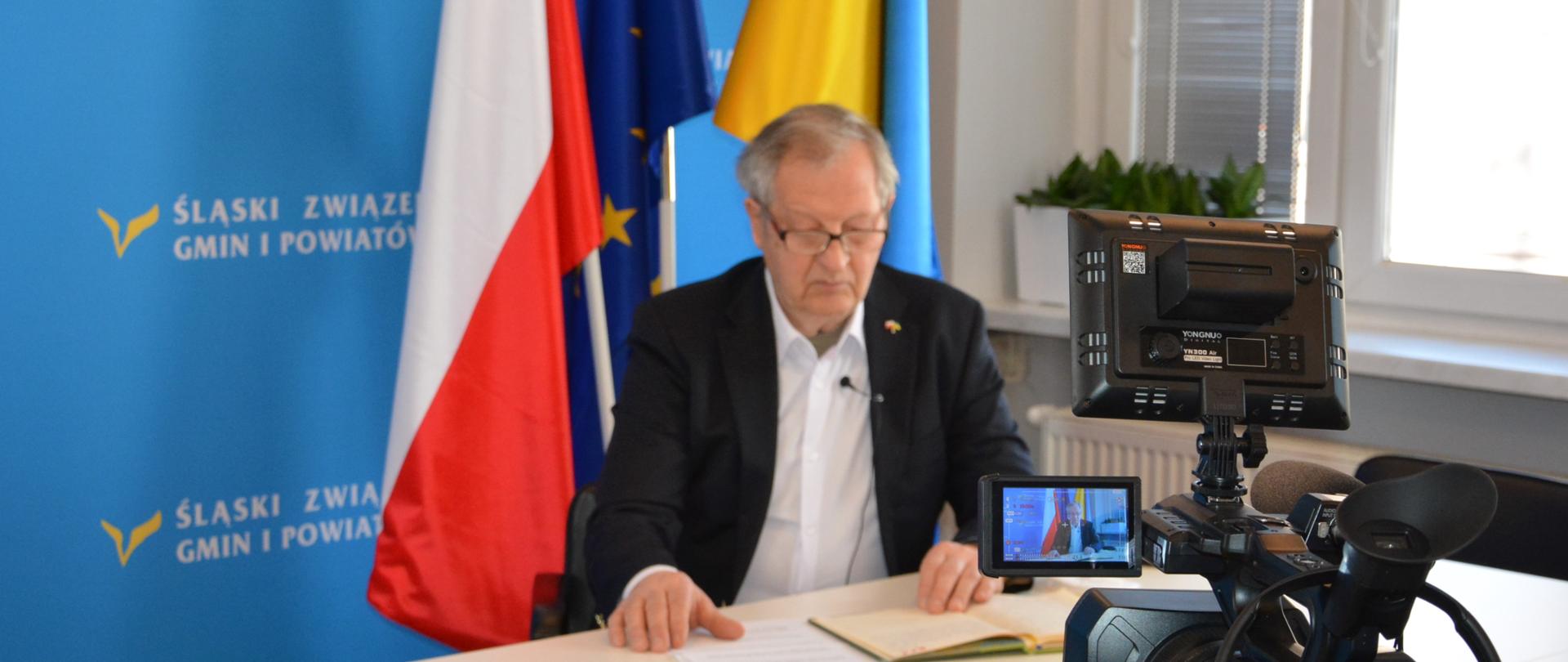 Mężczyzna (Garri Martin) siedzi przy stole i przegląda notes, kalendarz. Zdarzenie nagrywa widoczna na pierwszym planie kamera. w tle widoczna jest flaga Polski, Unii Europejskiej oraz Ukrainy.