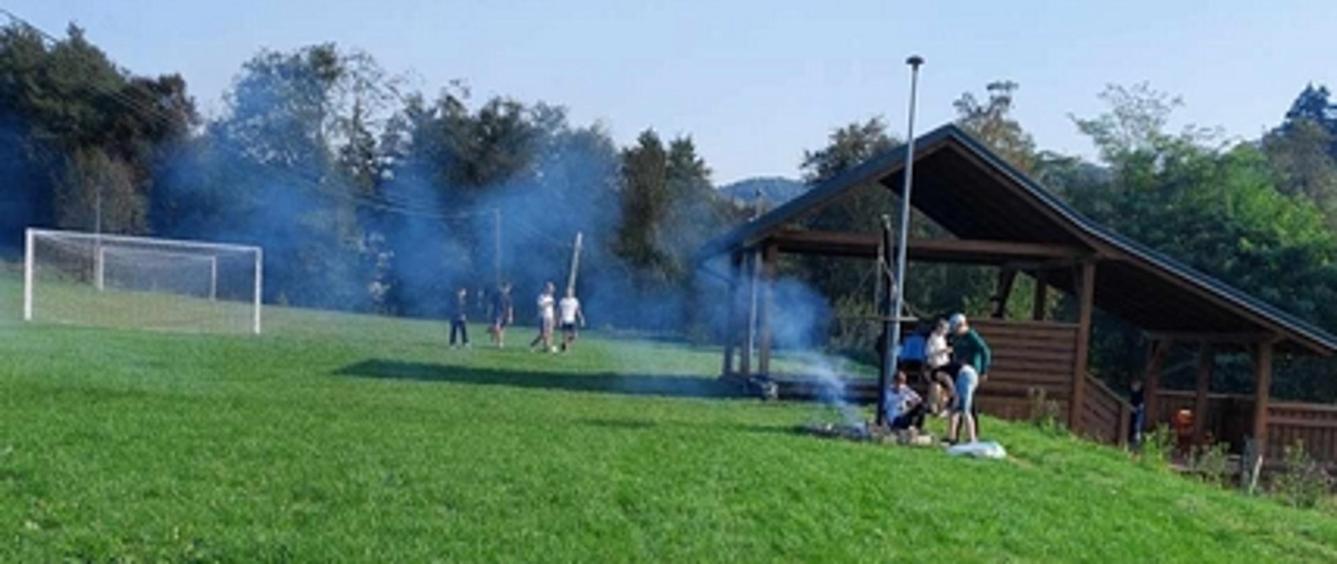 Pole z boiskiem do piłki nożnej. Grupka dzieci gra w piłkę nożną na boisku. Pozostałe dzieci siedzą przed drewnianą altanką przy rozpalonym ognisku